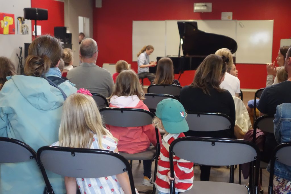 Des familles assises de dos en train d'écouter une jeune pianiste en fond.